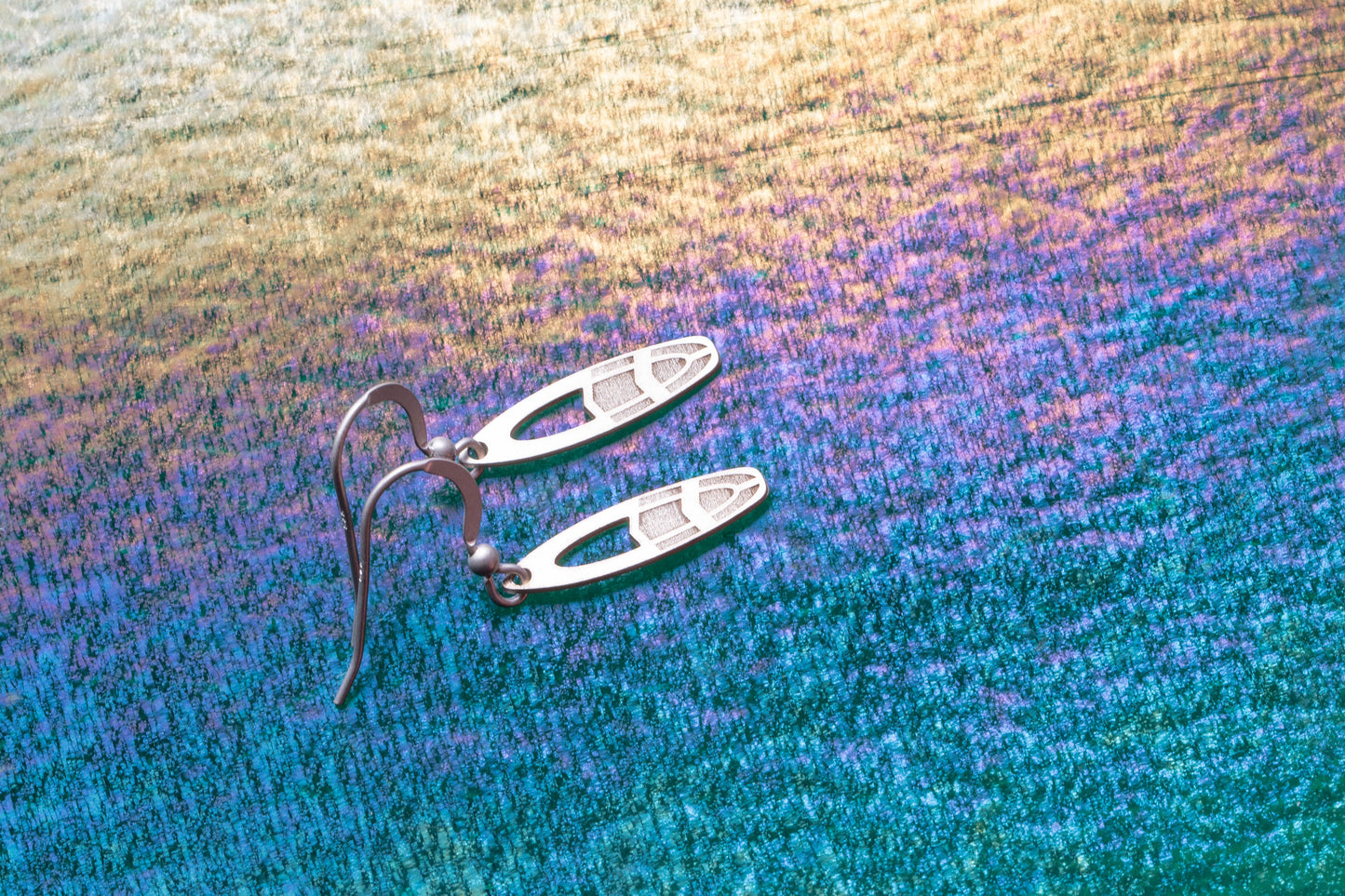 Small  fun surfboard shaped earrings shown on a blue and gold iridescent background